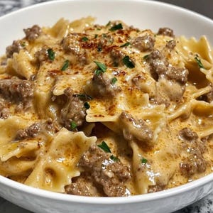 Op de afbeelding: Een witte kom gevuld met gekookte farfalle pasta, bedekt met een romige, lichtbruine saus. De saus is gemengd met gehakt en bestrooid met gehakte groene kruiden en een snufje rode kruiden. Close-up van een heerlijke maaltijd.