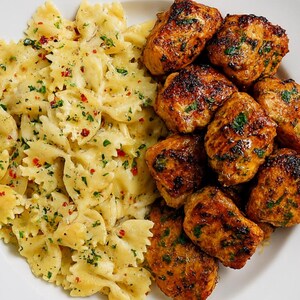 Pode incluir: Um prato de comida com massa farfalle em molho cremoso, salpicado com ervas e flocos de pimenta vermelha. Ao lado, vários pedaços de frango grelhado, com exterior dourado e ervas.