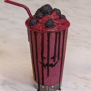 Op de afbeelding: Een hoog glas gevuld met een donkerpaarse smoothie, gegarneerd met verse bramen. Donkere siroop druipt langs de zijkanten van het glas en een griezelig pompoengezicht is op het glas getekend. Een rood rietje zit in de drank.
