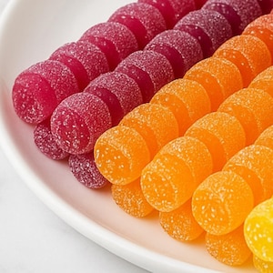 May include: Close-up of a white plate filled with colorful gummy candies. Rows of red, purple, orange, and yellow candies are arranged in diagonal lines. The candies have a sugared coating and a semi-circular shape.