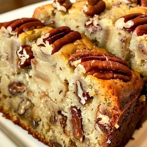 Könnte beinhalten: Nahaufnahme eines Stücks Pekannusskuchens. Der Kuchen hat eine hellbraune, strukturierte Oberfläche mit sichtbaren Pekannüssen oben und im Inneren. Die Pekannüsse haben eine satte, dunkelbraune Farbe. Der Kuchen liegt auf einem weißen Teller.