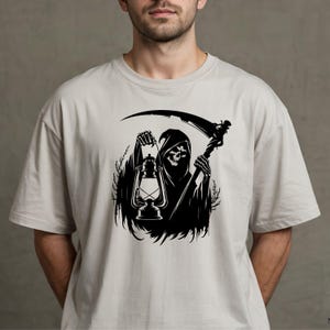 May include: A light gray t-shirt featuring a black graphic of the Grim Reaper holding a lantern and a scythe. The Grim Reaper is depicted in a hooded cloak with a skull face. The design is centered on the shirt.