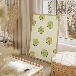 Peut inclure: Un cadre en bois clair présente une impression avec un motif de tranches de citron vert sur fond crème. L'œuvre est posée contre un mur. La pièce est décorée dans des tons neutres et bénéficie de la lumière naturelle.