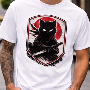 以下が含まれることがあります： 黒い猫の忍者のグラフィックが描かれた白いTシャツ。猫は忍者の衣装を着て、赤い太陽を背景に刀を持っています。デザインはシールド型のボーダーで囲まれています。
