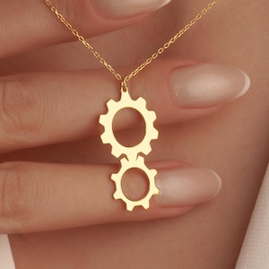May include: A gold-colored necklace featuring two interlocking gear pendants. The necklace is worn against a white top. The gears are detailed with circular cutouts. The chain is delicate and thin.