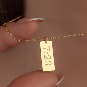 May include: A delicate gold necklace featuring a rectangular pendant. The pendant is engraved with the time "7:23". The necklace is displayed against a skin tone background, highlighting the jewelry's design and color.