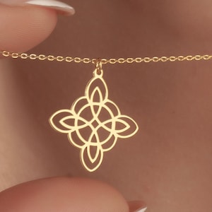 Könnte beinhalten: Eine goldfarbene Halskette mit einem keltischen Knotenanhänger. Der Anhänger ist ein symmetrisches, offenes Design, das an einer zarten Kette hängt. Die Halskette wird vor einem hautfarbenen Hintergrund präsentiert.