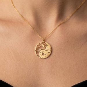 Könnte beinhalten: Eine goldfarbene Halskette mit einem runden Anhänger, der ein ausgeschnittenes Design einer aufgehenden Sonne über einem Gebirge und sanften Hügeln zeigt. Der Anhänger hängt an einer feinen goldfarbenen Kette am Schlüsselbein.