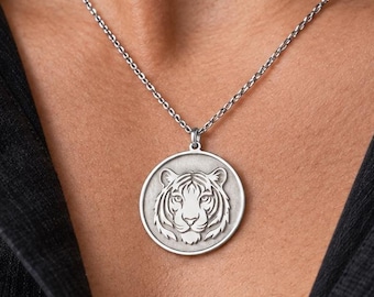 May include: A silver-toned circular pendant necklace featuring a detailed embossed tiger head design. The pendant hangs from a thin, matching silver-toned metal chain, resting against a dark gray collared shirt.