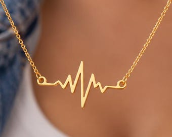 Könnte beinhalten: Eine goldfarbene Halskette mit einem zentralen Anhänger in Form einer EKG-Herzschlaglinie. Der Anhänger ist an einer zarten, dünnen goldfarbenen Kette befestigt, die an beiden Enden des Herzschlagmotivs ansetzt und ein durchgehendes Schmuckstück bildet.