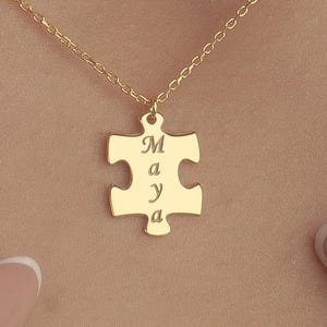 Puede incluir: Un collar dorado con un colgante de pieza de rompecabezas. El colgante tiene grabado el nombre "Maya". El collar se muestra sobre un fondo color piel.