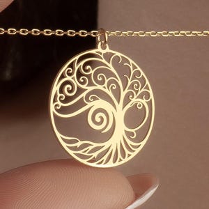 May include: Gold-colored necklace featuring a circular pendant with a tree of life design. The pendant has intricate, swirling details within the tree's branches and trunk. The necklace is held by a person's fingers, showcasing the delicate chain.