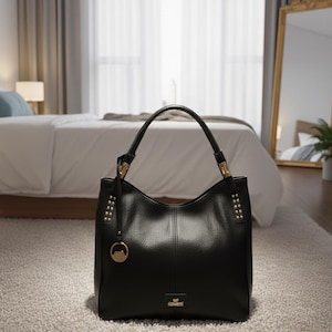 May include: Black leather tote bag with a structured design. The bag features a top handle, gold-toned hardware, and a decorative charm. The bag is sitting on a light-colored rug in a room with a bed and a mirror.