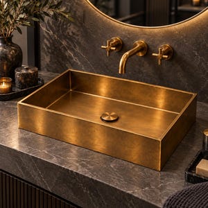 Peut inclure: Un lavabo rectangulaire en laiton avec une finition brossée et un bouchon de vidange circulaire assorti. Le lavabo est installé sur un comptoir en marbre gris foncé et est associé à un robinet mural en laiton avec deux poignées en croix.