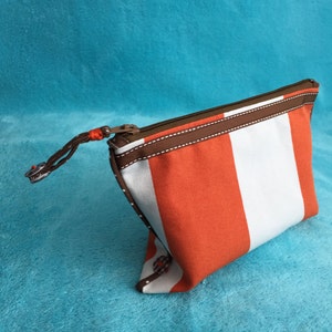 Könnte beinhalten: Kosmetiktasche aus orange-weiß gestreiftem Stoff mit braunem Reißverschluss und braunem Besatz.