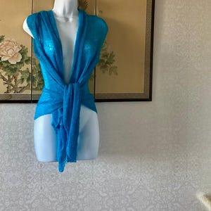 Könnte beinhalten: Ein leuchtend türkisblaues Wickeloberteil mit einer transparenten, leichten und strukturierten Strickkonstruktion. Das Kleidungsstück hat einen tiefen V-Ausschnitt und ist vorne gebunden, was einen drapierten Effekt auf dem weißen Schaufensterpuppen-Torso erzeugt.