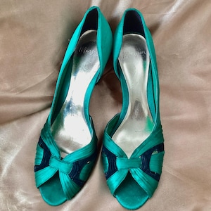 Könnte beinhalten: Ein Paar smaragdgrüne Satin-Pumps mit offener Zehenpartie und dunkelblauen Akzenten im Zehenbereich. Die Schuhe verfügen über ein silberfarbenes Innenfutter, wobei der Markenname Monsoon in Schwarz im Fersenbereich der Innensohle aufgedruckt ist.