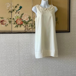 Könnte beinhalten: Cremefarbenes ärmelloses Kleid mit einem Bubikragen, der mit kleinen dekorativen Elementen verziert ist. Das Kleid hat eine einfache A-Linien-Silhouette und horizontale Streifen am Saum. Das Kleid wird auf einer Schaufensterpuppe präsentiert.