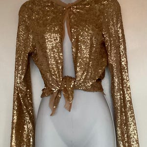 Könnte beinhalten: Goldfarbene Pailletten-Bolerojacke mit langen Ärmeln und Bindeband in der Taille. Die Jacke wird auf einer Schaufensterpuppe präsentiert und hat ein durchsichtiges, hautfarbenes Futter. Die Pailletten erzeugen einen schimmernden Effekt.
