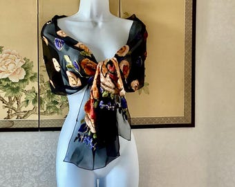 Puede incluir: Bufanda negra transparente con un estampado floral en tonos rojos, naranjas, azules y amarillos. La bufanda está drapeada sobre un maniquí, mostrando el diseño. El fondo presenta una pantalla decorativa con motivos florales y de aves.