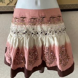 Könnte beinhalten: Ein rosa, weißer und brauner Rock mit floralen Stickereien und Pailletten-Akzenten. Der Rock hat ein mehrschichtiges Design mit Rüschensaum und Wickelstil. Der Rock wird auf einer Schaufensterpuppe präsentiert.