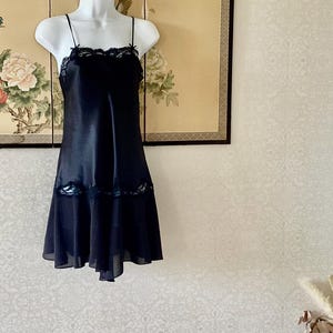 May include: A black slip dress featuring thin spaghetti straps, a straight neckline with black lace trim, and a matching lace panel detail across the mid-section. The garment has a flared skirt portion with a sheer hemline and a smooth finish.