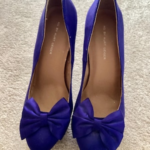 May include: A pair of royal blue high-heeled shoes with a satin finish. Each shoe features a large bow on the toe and a pointed toe design. The inner sole is a light brown color, and the brand name "KG by Kurt Geiger" is printed inside.