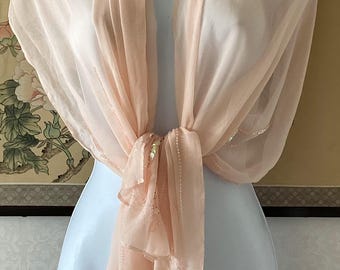 May include: A delicate, peach-coloured scarf draped over a mannequin. The scarf is knotted at the front and has subtle sequin detailing along the edges. The fabric is lightweight and flowing, suggesting a formal or elegant accessory.