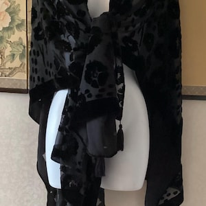 Puede incluir: Chal negro con estampado floral y borlas. El chal está drapeado sobre un maniquí blanco. La tela tiene una textura aterciopelada, con un fondo transparente. Las borlas cuelgan de los bordes inferiores.