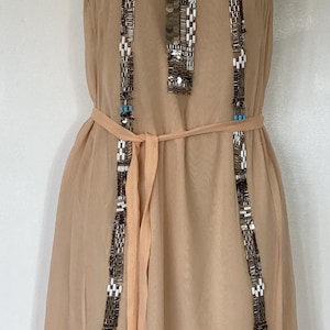 May include: A beige, sleeveless dress with intricate beaded embellishments. The dress features a halter neckline and vertical beaded stripes. A matching sash cinches the waist. The dress is displayed on a mannequin.