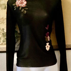 Könnte beinhalten: Schwarzes Langarm-Top mit Stehkragen. Der transparente Stoff ist mit gestickten Blumenmustern in Rosa, Rot und Grün verziert. Das Top hat eine figurbetonte Silhouette und wird auf einer Schaufensterpuppe präsentiert.