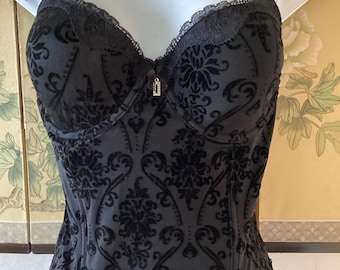May include: A black corset with a floral damask pattern. The corset has adjustable straps, a lace trim, and a small decorative charm. The fabric appears to be a velvet-like material. The corset is displayed on a white mannequin.
