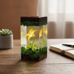 May include: A rectangular resin art piece featuring a yellow lily flower encased within clear resin. The base of the piece has green moss and a dark brown border. The lily has green leaves and a yellow flower.