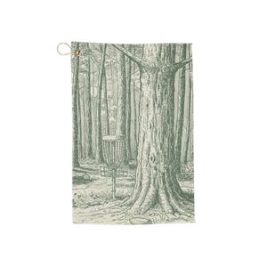 May include: A green and white disc golf towel depicting a forest scene with a disc golf basket. The towel has a brass grommet for hanging. The design is a detailed illustration of a disc golf course.