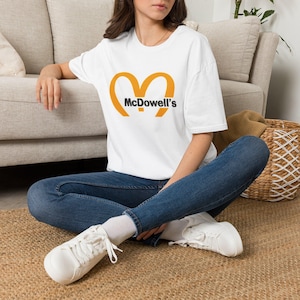May include: A white short-sleeved t-shirt featuring a large yellow graphic of two curved arches forming an M shape with the black text McDowell's printed across the center. The shirt is paired with dark blue denim jeans and white sneakers.