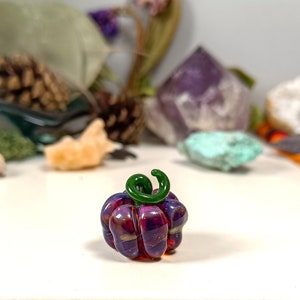 May include: A small glass pumpkin with a green stem. The pumpkin is a deep purple colour with swirls of gold and red. It is sitting on a white surface.