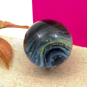 Pode incluir: Uma esfera de vidro com um design azul, verde e dourado em espiral. A esfera est&aacute; cheia de pequenas manchas douradas, criando um efeito de gal&aacute;xia.