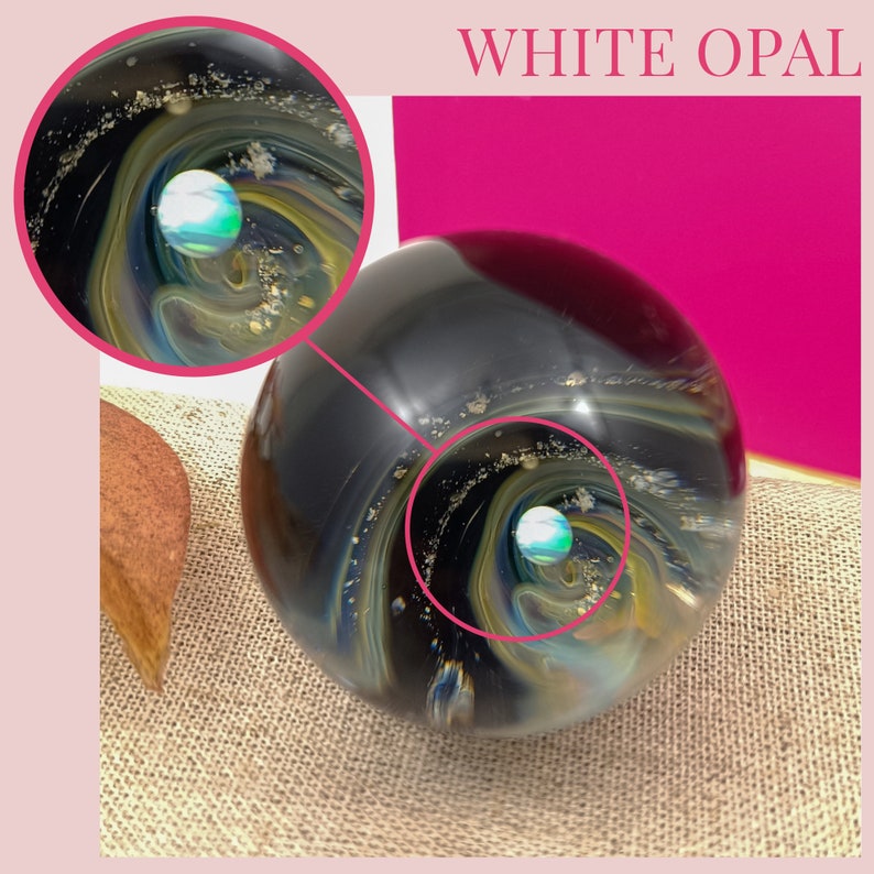 Pode incluir: Uma bola de vidro de opala branca com um design em espiral preto, azul e dourado. A bola tem um pequeno orbe redondo azul e verde no centro.