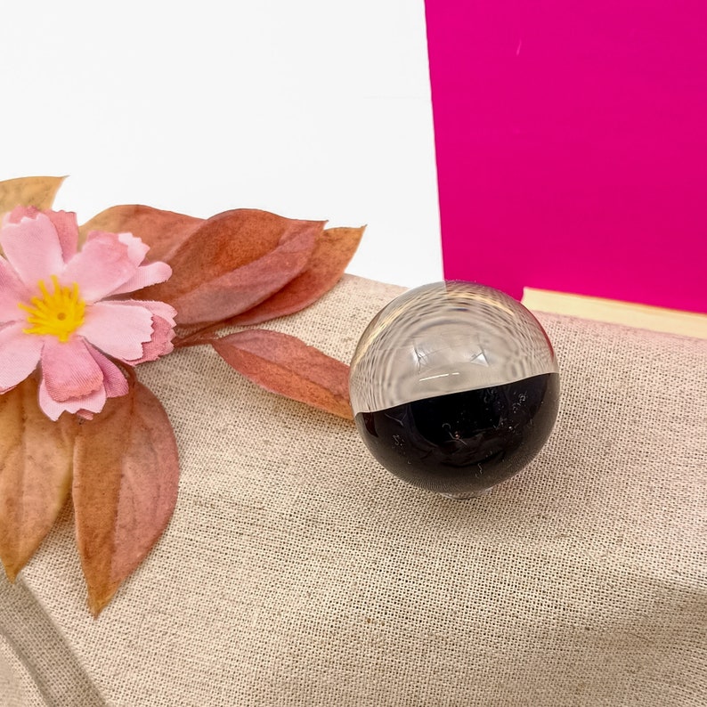 Pode incluir: Uma esfera de vidro transparente com metade inferior preta. A esfera est&aacute; sobre uma superf&iacute;cie de tecido bege.