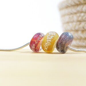 Puede incluir: Tres cuentas de vidrio en una cadena plateada. Las cuentas son de color rojo oscuro, amarillo claro y gris oscuro. Cada cuenta tiene un patr&oacute;n &uacute;nico de burbujas y remolinos.