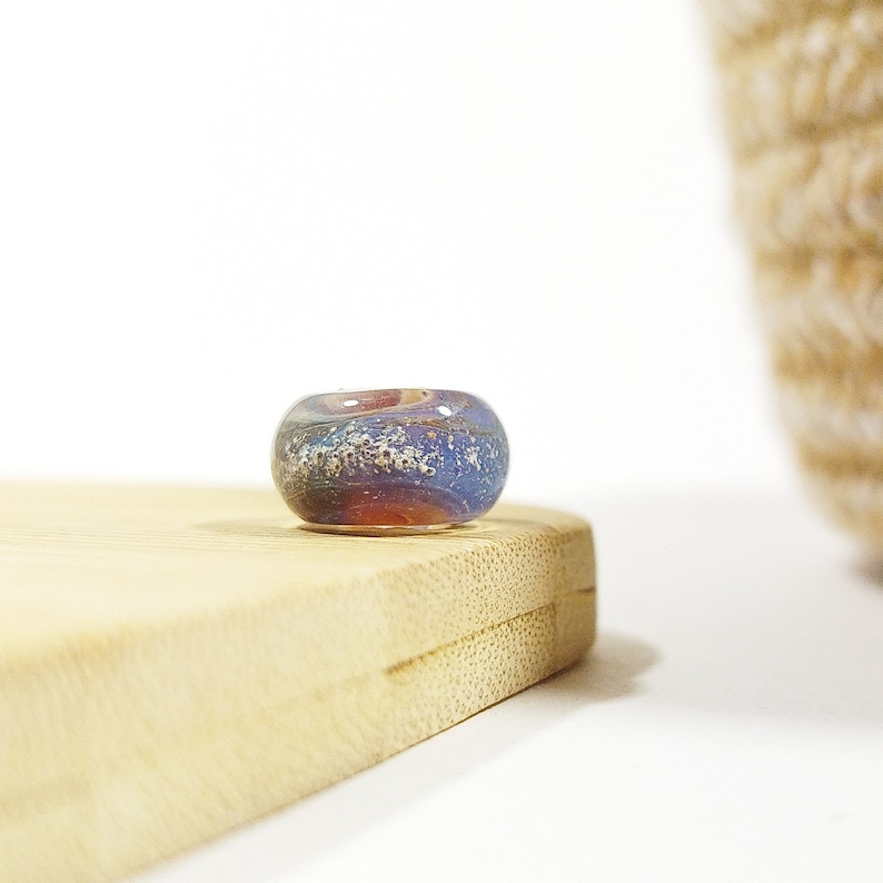 Puede incluir: Una cuenta de vidrio azul, p&uacute;rpura y dorada con un patr&oacute;n de remolino. La cuenta est&aacute; sobre una superficie de madera.