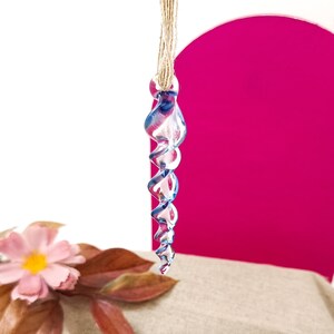 May include: A blue glass icicle ornament with a spiral design hangs from a twine string. The ornament is suspended in front of a pink background.