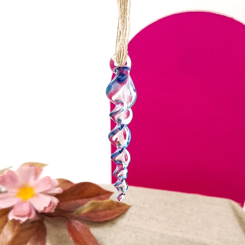 May include: A blue glass icicle ornament with a spiral design hangs from a twine string. The ornament is suspended in front of a pink background.