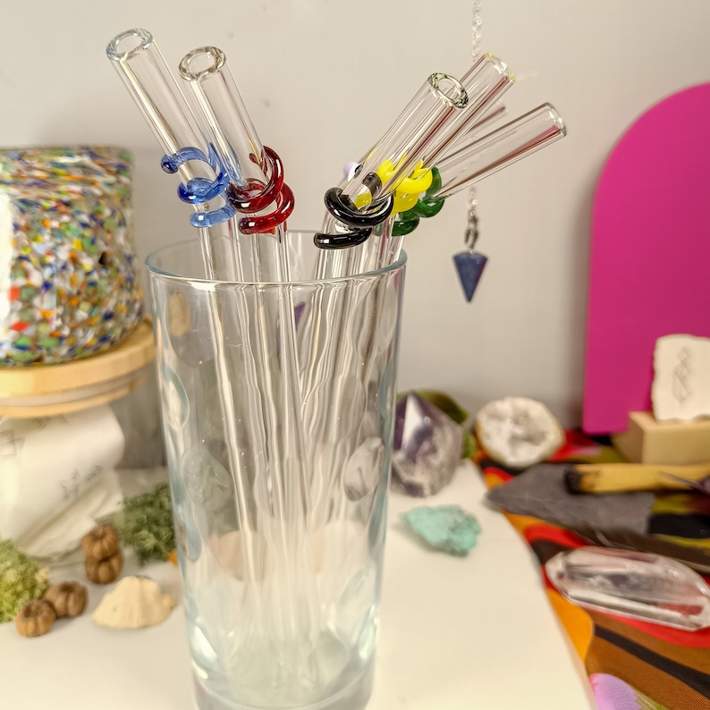 May include: A set of six glass drinking straws with colorful silicone tips. The straws are in a clear glass cup on a table with various crystals and other decorative items.
