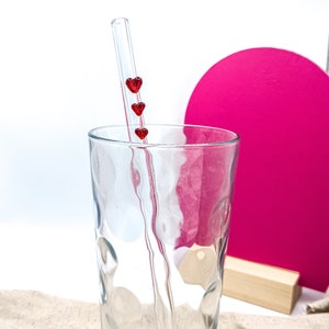 May include: A clear glass drinking straw with three red heart-shaped charms. The straw is in a clear glass with a textured surface. The glass is on a light brown surface.