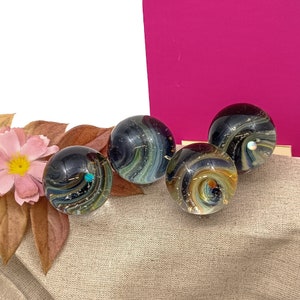 Pode incluir: Quatro bolas de vidro com padr&otilde;es em espiral de preto, branco e dourado. As bolas est&atilde;o dispostas sobre uma superf&iacute;cie de tecido bege.