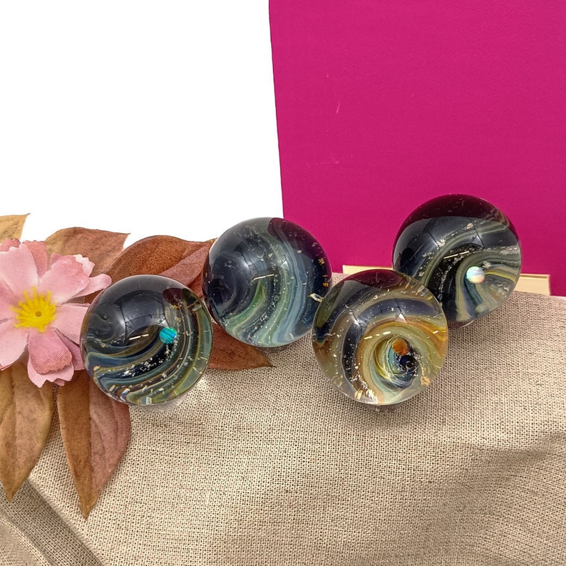 Pode incluir: Quatro bolas de vidro com padr&otilde;es em espiral de preto, branco e dourado. As bolas est&atilde;o dispostas sobre uma superf&iacute;cie de tecido bege.