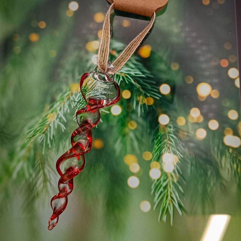 May include: A red glass Christmas bauble shaped like a spiral. The bauble is hanging from a twine string against a blurred background of a green Christmas tree.