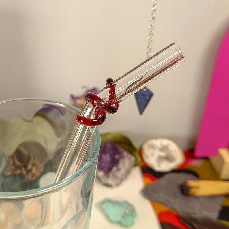 May include: A clear glass drinking straw with a red glass swirl design. The straw is in a glass with a clear liquid.