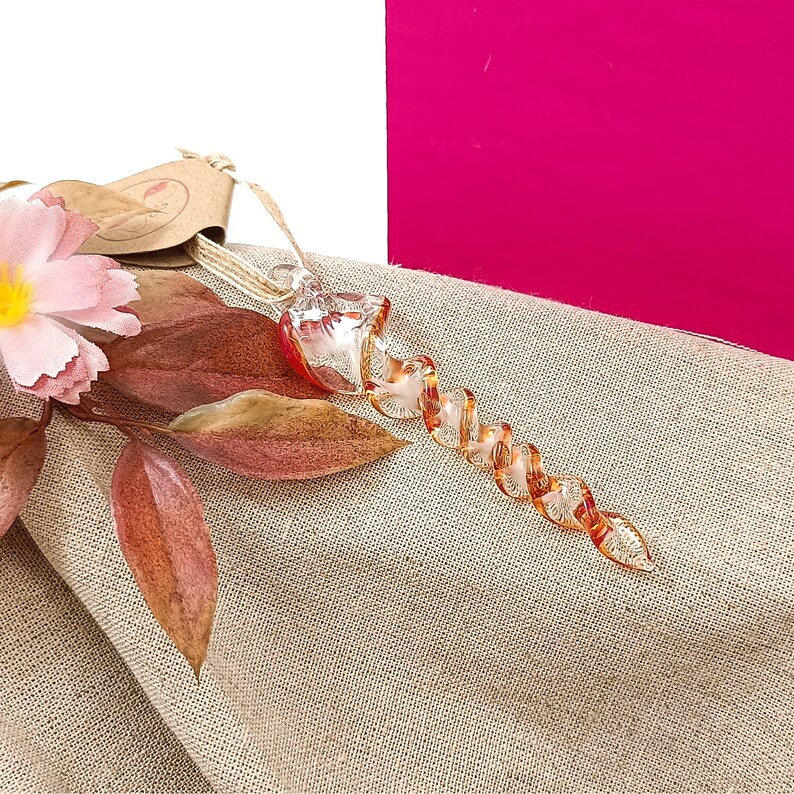 May include: A glass ornament shaped like a spiral with a clear and amber colour scheme. The ornament is lying on a beige fabric surface with a pink flower and brown leaves.
